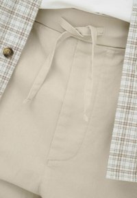 Pantalon beige à cordon de serrage associé à une veste beige et marron à carreaux avec un bouton marron.