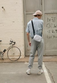 Un pantalon de jogging gris, une chemise légère à manches courtes bleu clair, une casquette beige et un petit sac bandoulière blanc. Un vélo avec des pneus fins est à proximité.