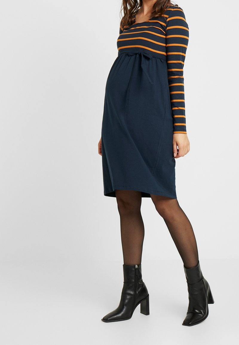 Robe de maternité avec corsage bleu marine et rayures horizontales orange. Dotée d'une taille froncée avec un détail de nœud. Bottines noires complètent le look.