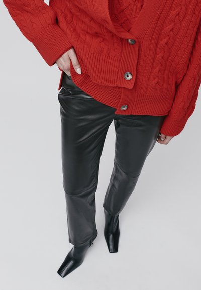 Raudonas megztinis su iškeltomis akytėmis ir mygtukais, derinamas su juodomis odinėmis kelnėmis ir smailiais juodais batais. Akcentuojamas tekstūros ir spalvų kontrastas.