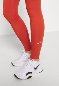Czerwone legginsy sportowe o gładkiej fakturze z białym logo Nike, zestawione z białymi butami Nike z czarnymi akcentami.