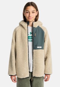 Giacca in pile beige con cappuccio, caratterizzata da un pannello in nylon verde sul davanti con cerniera e un piccolo tag di branding, indossata sopra una maglietta grigia.