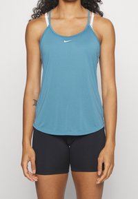 Top sportivo blu con spalline bianche sottili, orlo curvo e un piccolo logo Nike sul davanti. Abbinato a pantaloni corti aderenti neri.