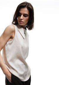 Ärmlös beige blus med spetsig krage, prydd med en rund knappbetoning vid halsen och en mjuk, sidenliknande textur.