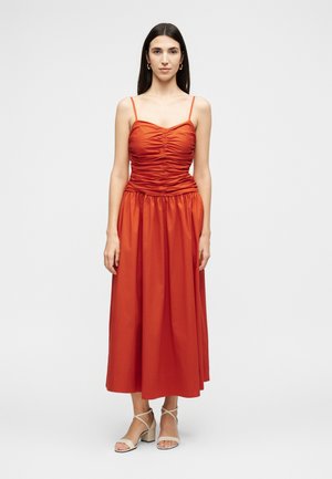 Kobieta z prostymi ciemnymi włosami, ubrana w pomarańczową marszczoną sukienkę midi i beżowe sandały na obcasie, stojąca na tle jednolitego jasnoszarego tła.