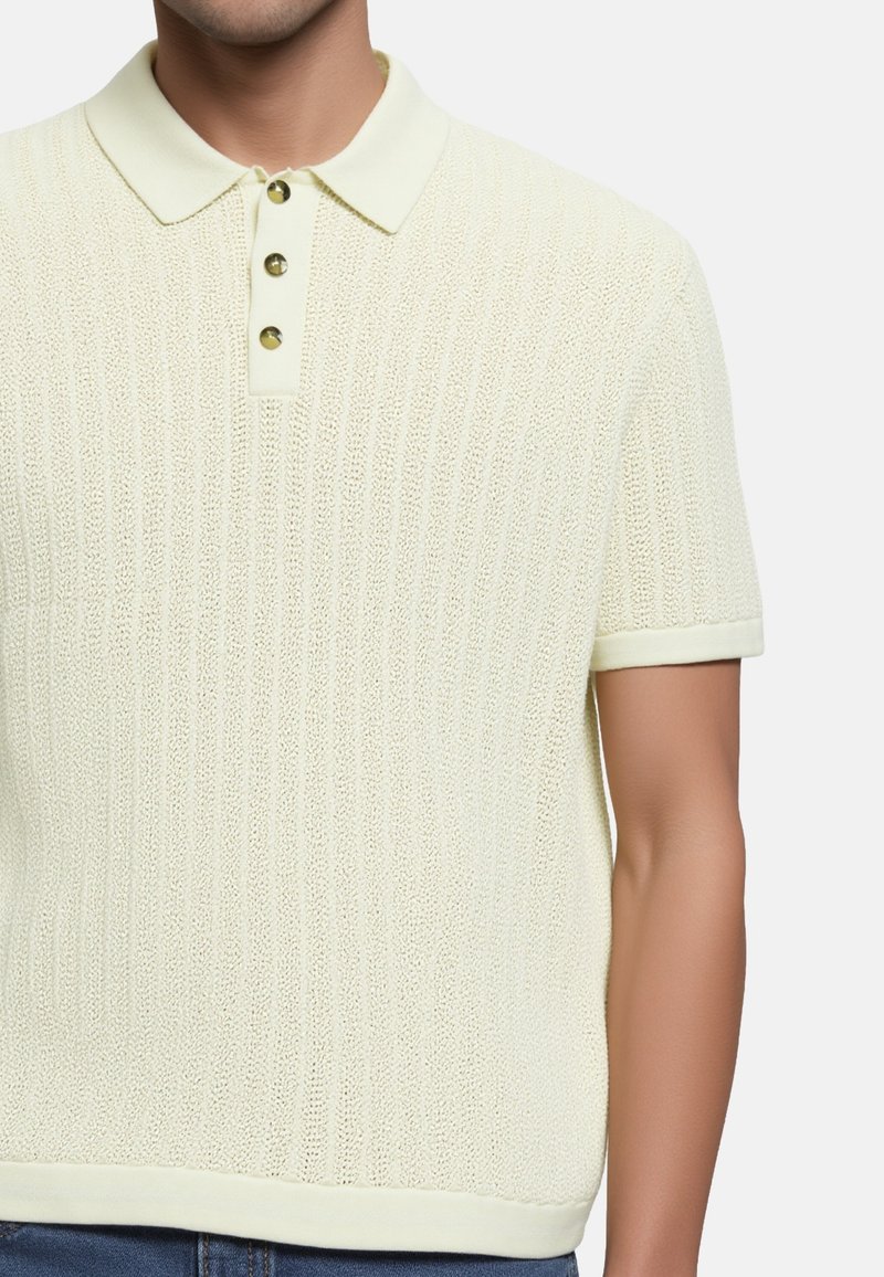 Hombre que lleva una camiseta polo de manga corta texturizada en color crema con tres botones, combinada con jeans azules, frente a un fondo liso.