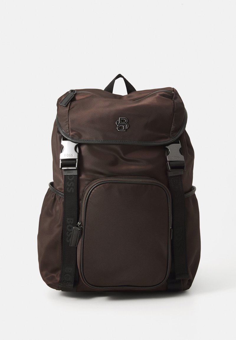 Mochila castanho escura com bolso frontal com fecho, dois bolsos laterais, alças ajustáveis pretas com fivelas de metal e uma pega superior, sobre um fundo branco.