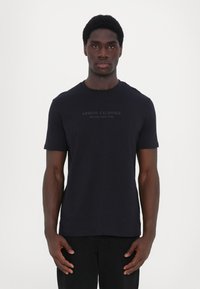 Juodas trumpomis rankovėmis marškinėliai, pagaminti iš minkštos medvilnės, su apvaliu iškirpimu ir spausdintu logotipu "Armani Exchange Milano/New York" priekyje.
