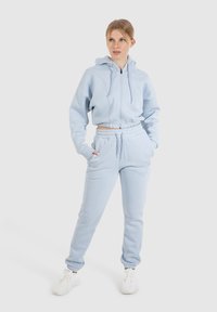 Felpa cropped azzurra chiara con zip, tasche a scomparsa e coulisse. Pantaloni jogger coordinati con vita ed elastico alle caviglie. Tessuto morbido.