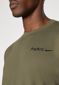 Olive zelené bavlnené tričko s okrúhlym výstrihom, zdobené čiernym vyšitým textom "PARÍŽ" a logom Nike. Hladká textúra a priliehavý strih.