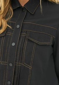 Sort skjorte med krave, der har kontrastfarvet gul syning, to brystlommer og trykknapper foran.