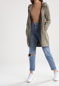 Olivově zelená bunda s kapucí a zipem, nošená přes béžový rolák. Kombinováno se světle modrými roztrhanými džínami a bílými teniskami.