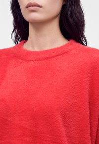 Femme aux longs cheveux foncés portant un pull rouge vif et duveteux, montrant le cou et le bas du visage sur un fond uni.
