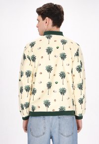 Lichtgele bomberjack met groene kraag, versierd met een verspreid patroon van palmbomen, avocado's en kokosnoten. Zachte, gladde textuur.