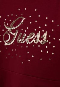 Tessuto borgogna con paillettes argento che formano la parola "Guess," circondata da paillettes argento sparse. Texture liscia con un design aderente.