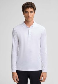 Polo shirt bianco a maniche lunghe con tessuto texturizzato, caratterizzato da un colletto piatto e una patta con tre bottoni. Abbinato a pantaloni scuri.