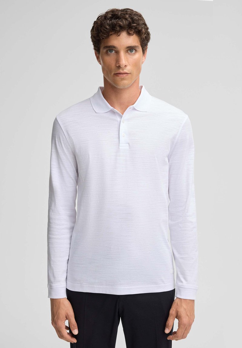 Polo shirt bianco a maniche lunghe con tessuto texturizzato, caratterizzato da un colletto piatto e una patta con tre bottoni. Abbinato a pantaloni scuri.