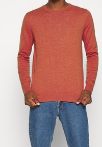 Pull rouge en maille avec manches longues et col rond. Comprend des poignets et un ourlet côtelés. Porté avec un jean denim bleu.