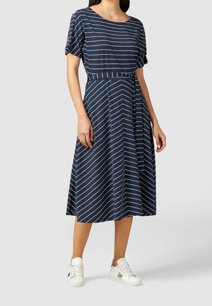 Femme portant une robe midi bleu marine avec des rayures diagonales blanches, manches courtes, ceinture à la taille et baskets blanches sur un fond gris clair.