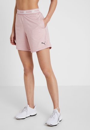 Vrouw gekleed in lichtroze Puma sportshorts en witte sneakers, staande met één hand in de zak tegen een effen achtergrond.