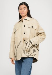 Giacca beige con bottoni neri e due tasche sul petto, caratterizzata da ricami floreali lungo il fondo. Tessuto morbido e vestibilità rilassata.