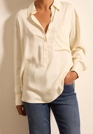 Osoba nosząca luźną, kremową, długą bluzkę z guzikami i przednią kieszenią, w połączeniu z niebieskimi dżinsami, ręka w kieszeni.