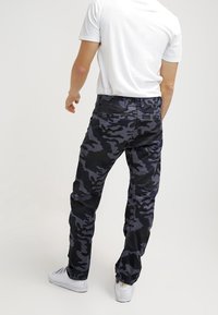 Man med mörkblå och svarta kamouflagebyxor, vit t-shirt och vita sneakers, stående vänd bort från kameran på en enfärgad bakgrund.