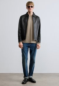 Fekete bőrdzseki galléros nyakkal, bézs hosszú ujjú ing, kék farmernadrág és fekete cipő. Kiegészítők között sötét napszemüveg található.