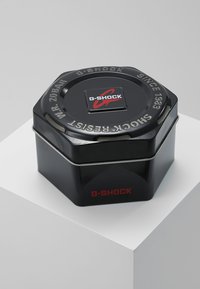 Conteneur en métal noir hexagonal avec une finition brillante, arborant un logo G-SHOCK en relief et des accents rouges, texte en relief sur le couvercle.