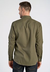 Camisa verde de manga larga con cuello, puños abotonados y una textura suave. Presenta una espalda con pliegues y está metida dentro de unos jeans de mezclilla oscura.
