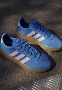 Pair of blue Adidas sneakers with white stripes and gum soles on concrete ground.