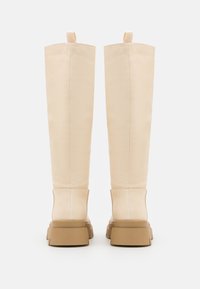 Botas de couro bege até ao joelho com uma textura suave, solas de borracha castanha robustas e alças de tecido no topo para um fácil calçar.