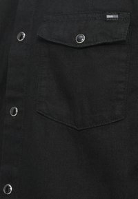 Juodas džinsinis marškinis su sidabriniais spaudimo sagomis ir krūtinės kišene su skydeliu. Medžiaga tekstūruota, su matinio paviršiaus apdaila ir tvarkingais siūlais.