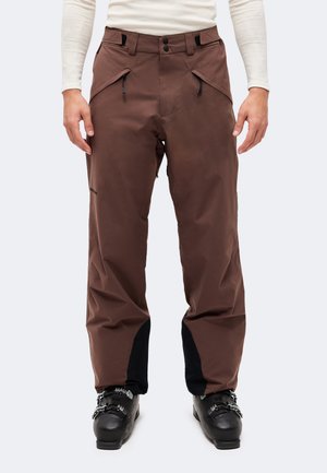 Modèle portant un pantalon de ski marron avec des poches zippées et des chaussures de ski noires, sur un fond clair uni.