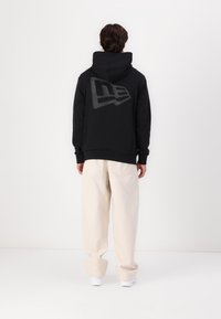 Felpa nera con un grande logo grigio sul retro, abbinata a pantaloni larghi beige. Il tessuto sembra liscio, con una vestibilità comoda.