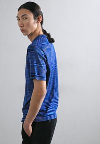 Camisa polo de mangas curtas azul riscada com colarinho, apresentando várias tonalidades de azul. Usada com calças pretas texturizadas.