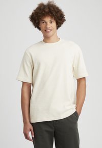 T-shirt à manches courtes, col rond en tissu crème texturé, présentant une coupe décontractée et un design simple, associé à un pantalon vert foncé.