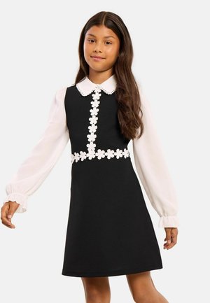 Girl wearing black sleeveless dress with white floral trim over a long-sleeve white blouse with a scalloped collar and ruffled cuffs.