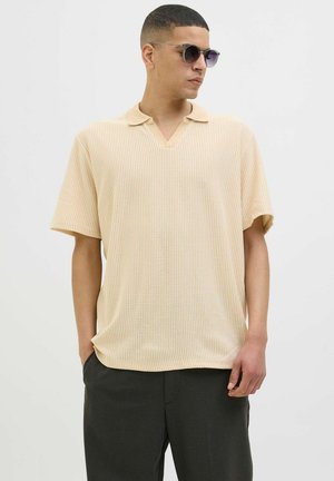 Chemise à manches courtes, col en V de couleur beige clair avec des rayures verticales, dotée d'une texture douce et d'une coupe décontractée, associée à un pantalon sombre et des lunettes de soleil.