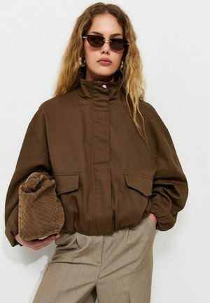 Woman wearing brown oversized jacket and beige pants, holding a textured brown clutch, with sunglasses and long wavy hair.
