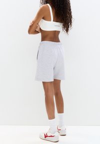 Reggiseno sportivo bianco con nome del marchio, pantaloni corti grigi in cotone con vita elastica e scarpe da ginnastica bianche con dettagli rossi e calzini bianchi.