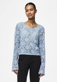 Light blue lace long-sleeve top with floral pattern, round neckline, and flared sleeves, worn over black pants.