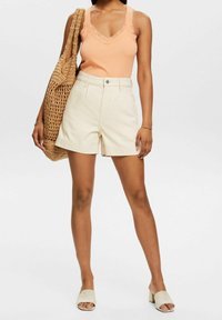 Top de tirantes de melocotón con encaje, pantalones cortos de talle alto en beige claro y sandalias de tacón bloque en beige; acompañado de un bolso de lona tejido.