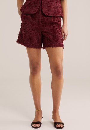 Nő bordó, texturált rövidnadrágot és hozzáillő mellényt visel, fekete nyitott orrú szandált és arany bokaláncot, egyszerű háttér előtt áll.