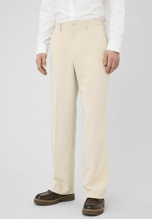 Homme portant un pantalon beige sur mesure, une chemise blanche et des chaussures en cuir marron, debout devant un fond clair uni.