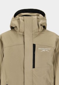 Beige Softshell-Jacke mit hohem Kragen, Reißverschlusstaschen und schwarzem Innenfutter. Mit weißem Logo-Detail und minimalistischer Gestaltung. Wasserdichtes Material.