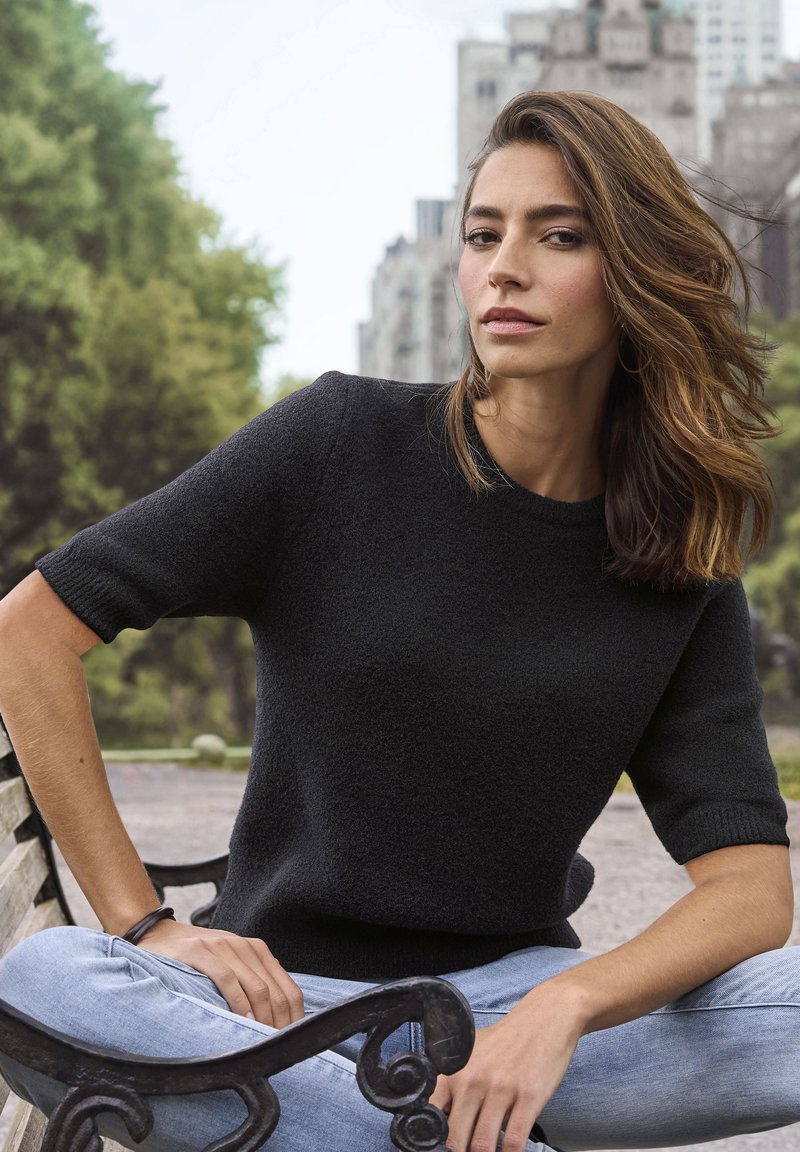 Čierny krátky rukávový pletený sveter s textúrovým povrchom, okrúhlym výstrihom a rebrovanými manžetami, v kombinácii so svetlomodrými džínsami, sedí na lavičke.