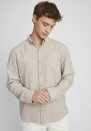 Chemise à boutons beige avec un motif texturé, manches longues, une poche poitrine et col boutonné, portée sur un pantalon blanc.