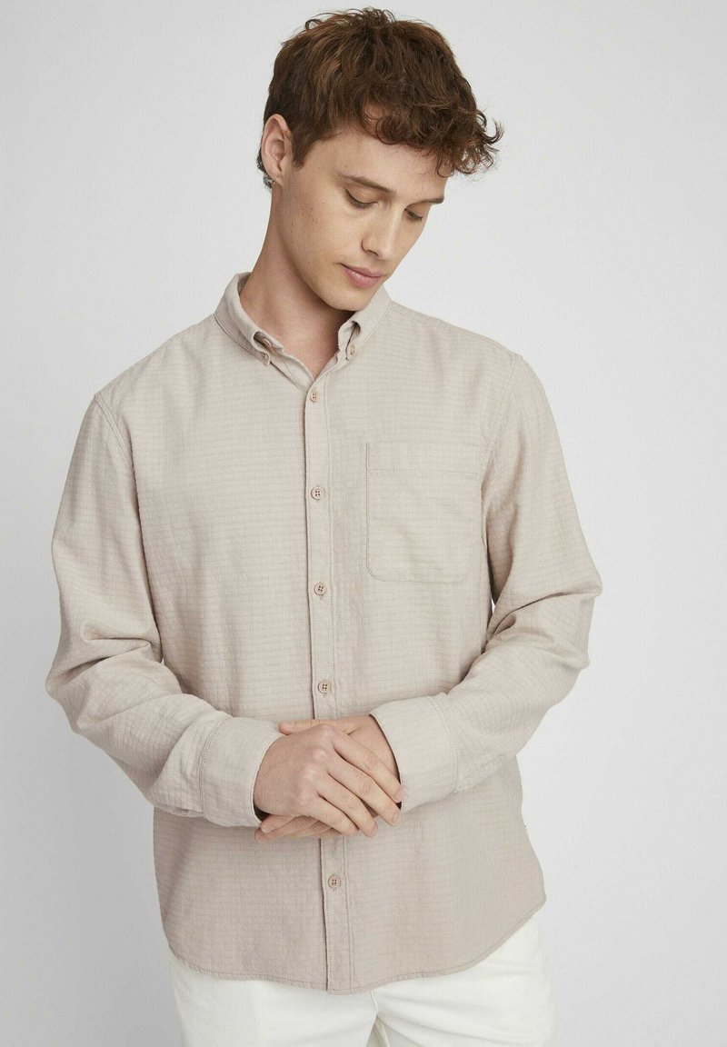 Chemise à boutons beige avec un motif texturé, manches longues, une poche poitrine et col boutonné, portée sur un pantalon blanc.