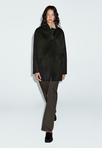 Zwarte wollen jas met een minimalistisch ontwerp, voorzien van een enkele knoopsluiting, met een ontspannen pasvorm en gecombineerd met olijfgroene broek en zwarte schoenen.